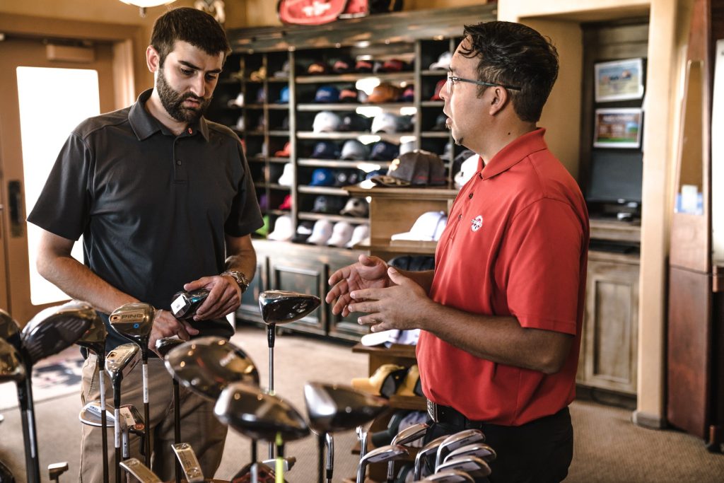 The person on the left is wearing a black polo shirt and is holding a golf club. The person on the right is wearing a red polo shirt and is gesturing with his hands. The background consists of a wooden display case with golf hats and a television screen.